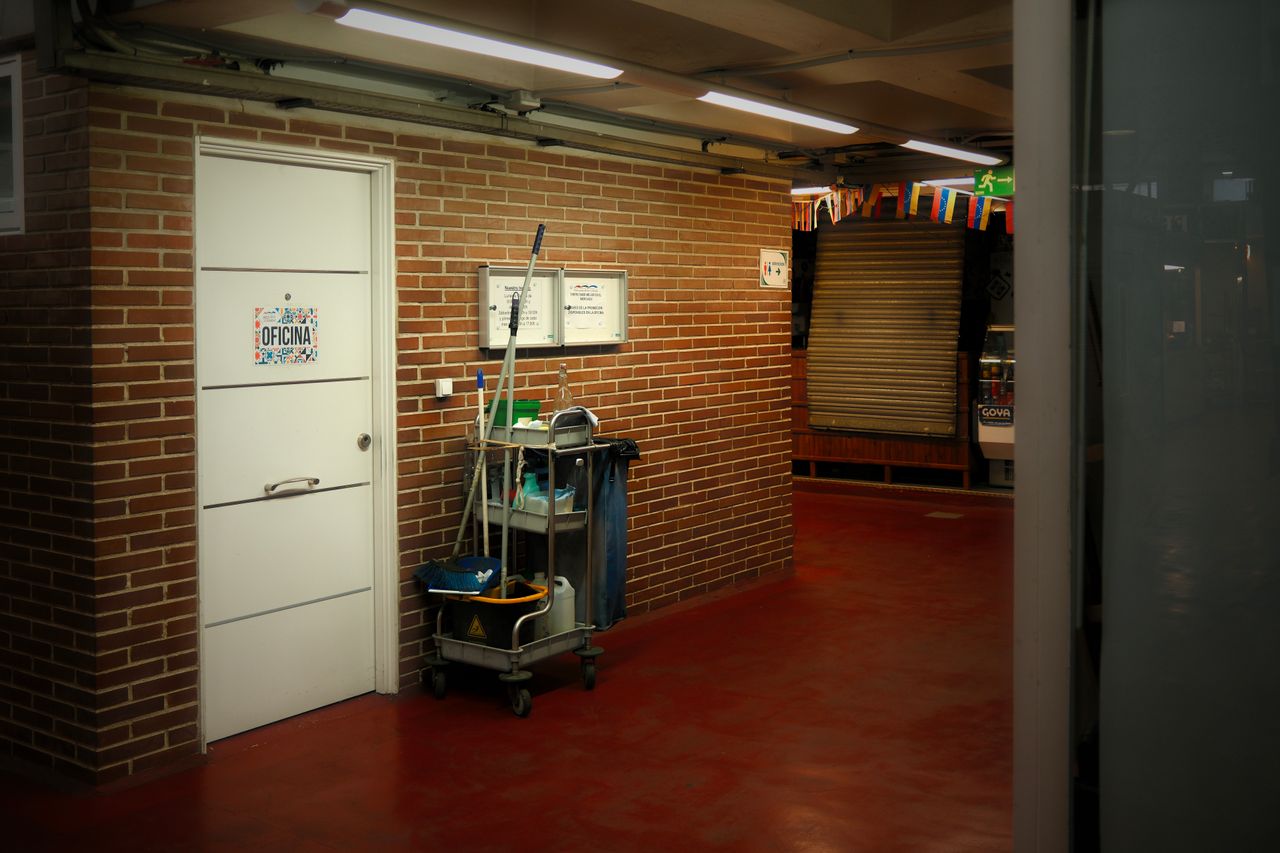 Un couloir industriel bien éclairé, avec un chariot de nettoyage et des fournitures près d’une porte de bureau.